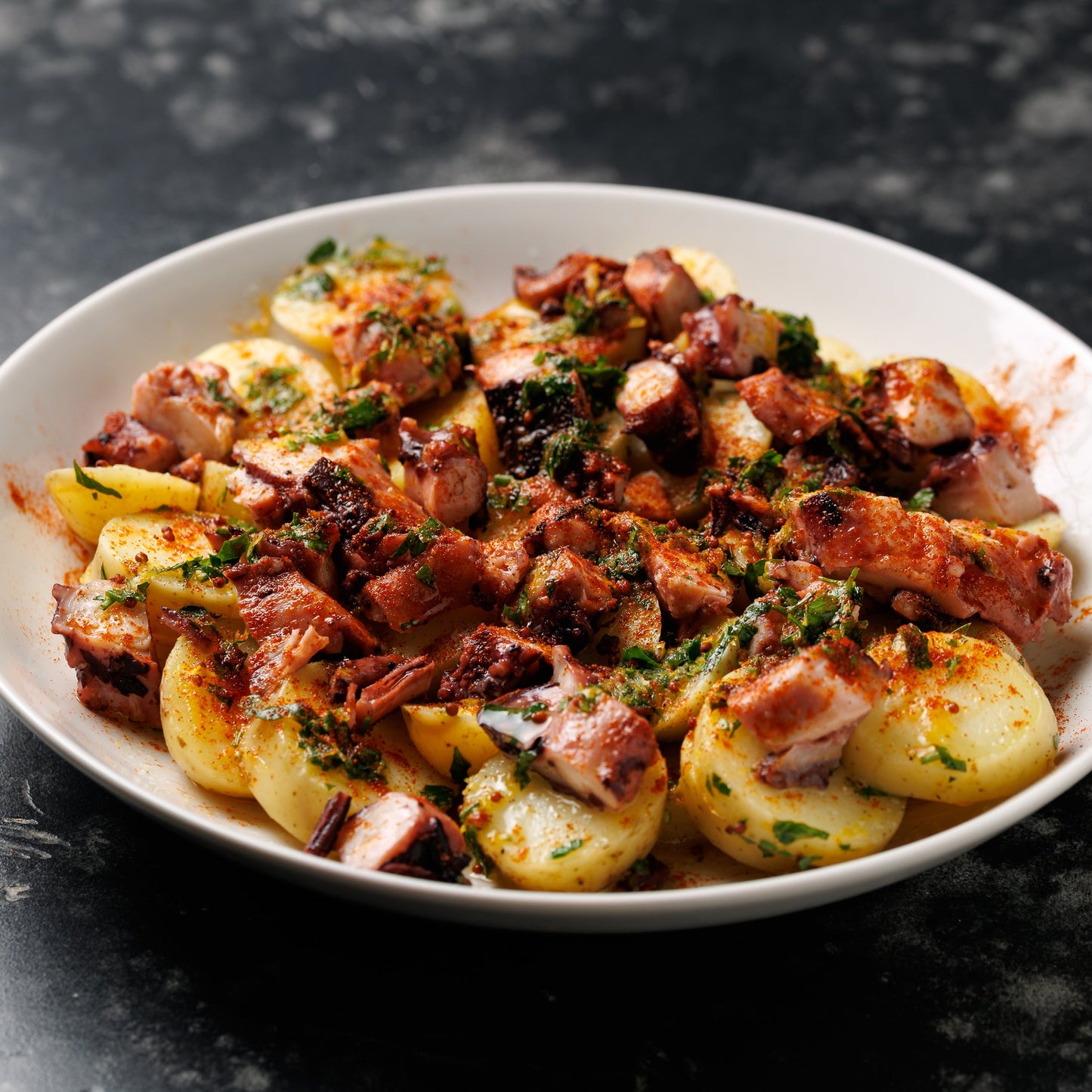 A white ceramic bowl filled with cooked potato slices, cubed sausage or bacon pieces, and a sprinkling of chopped green herbs, with a smoked paprika dressing set on a dark textured surface.