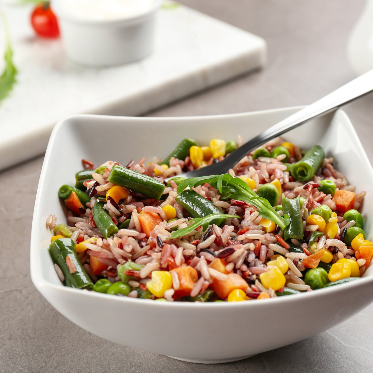 A colourful brown rice salad with mixed vegetables, leafy greens and fresh herbs served in a white bowl with a spoon, featuring Australian Brown Medium Grain Rice from The Herb Shed Wollongong Australia.