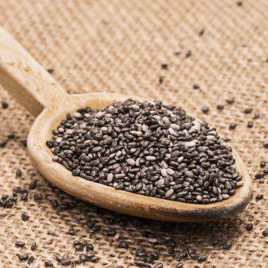 Black Chia Seeds fill a wooden spoon with loose seeds scattered across a natural hessian surface, showing their small oval shape and mottled colouring.