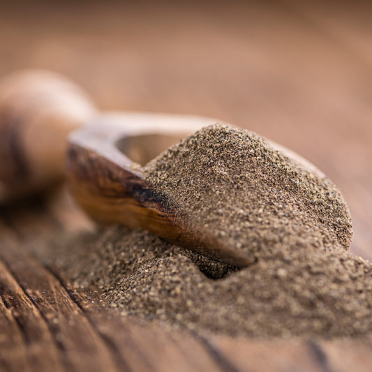 Ground black pepper fills a small wooden scoop on a rustic wooden surface, showing its finely ground texture and natural dark speckled colouring.