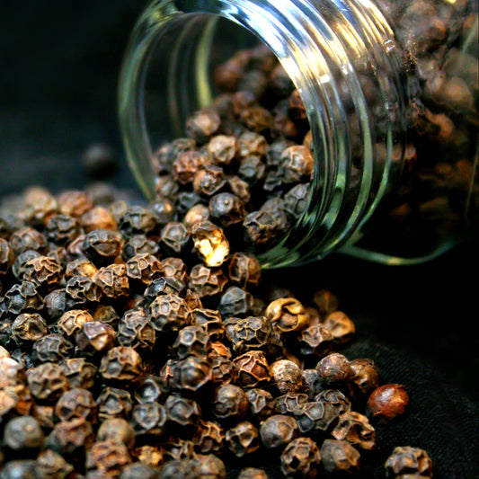 Whole black peppercorns spill from a glass jar onto a dark surface, showing their naturally wrinkled texture and varied dark brown and black colouring.