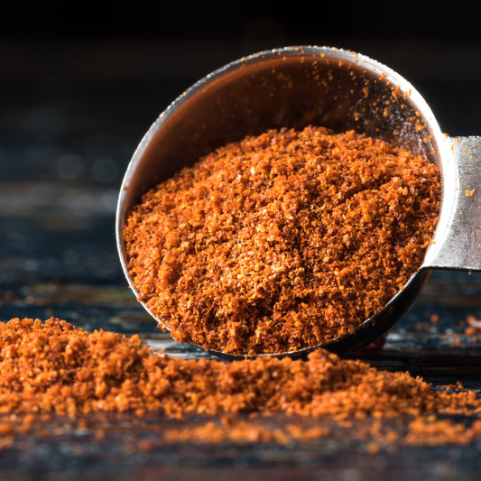 Ground Cayenne Pepper spills from a small bowl onto a dark rustic surface, showing its finely ground texture and rich red-orange colouring.