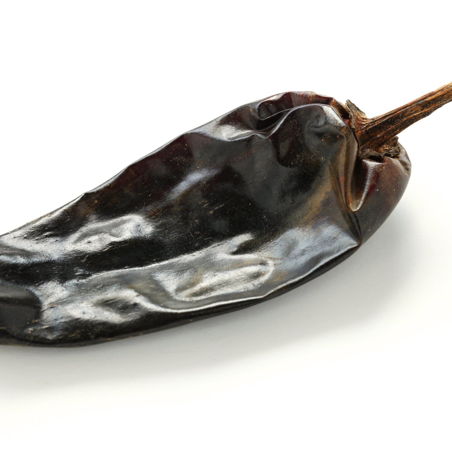 A single whole dried guajillo chilli is shown against a white background, displaying its dark red-brown colour, wrinkled skin and tapered shape.