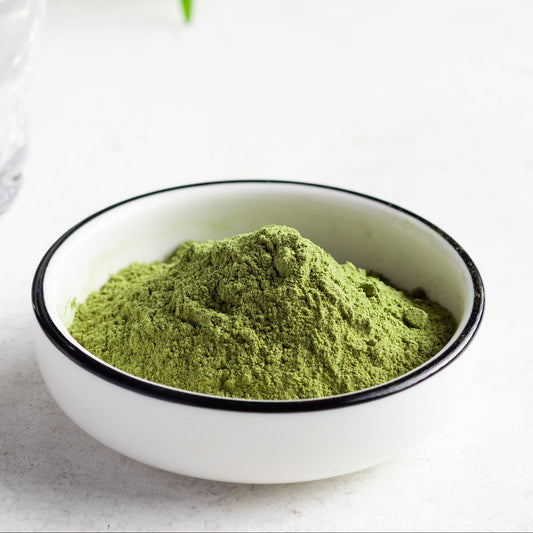Fine green lemongrass powder in a white-rimmed bowl beside a clear glass of water and a gold spoon on a light background – The Herb Shed Wollongong.