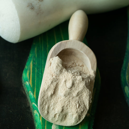 Fine onion powder fills a small white scoop resting on a green leaf, showing the pale cream colour and finely ground texture of the powder.