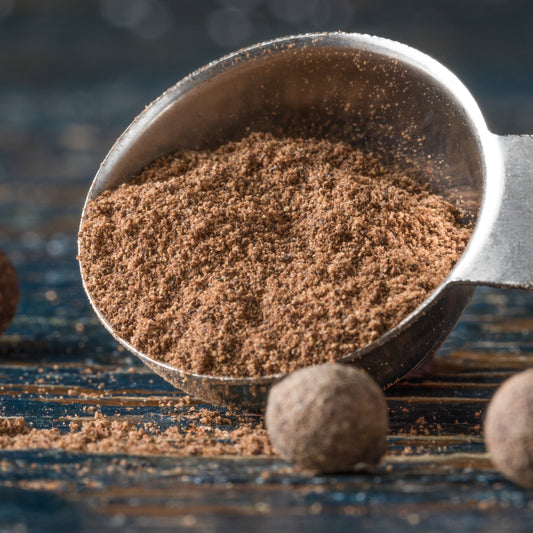 Ground Organic Allspice fills a metal scoop on a rustic wooden surface, with whole allspice berries scattered nearby.