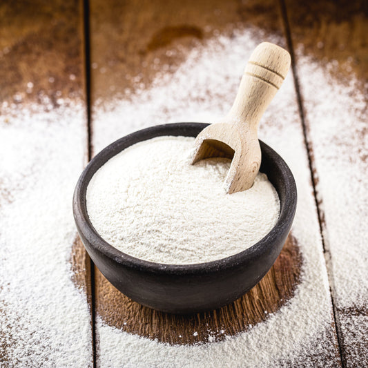 Organic Cassava Flour Extra Fine fills a dark bowl with a wooden scoop, showing its finely milled white texture against a rustic wooden surface.