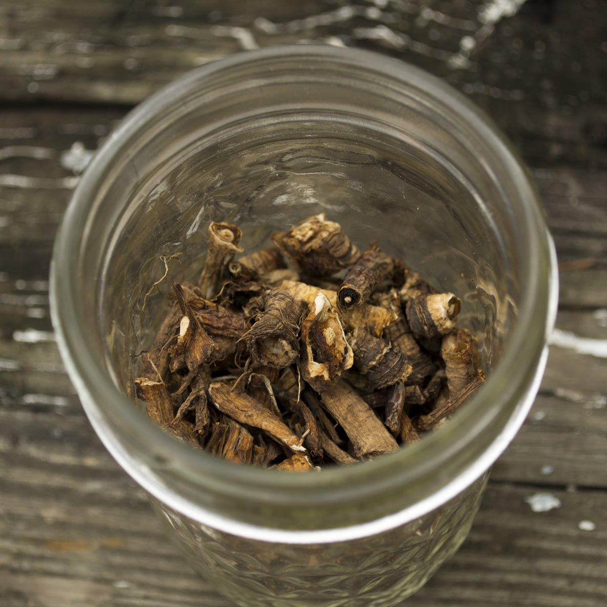 Roasted organic dandelion root pieces sit inside a clear glass jar on a rustic wooden surface, showing their irregular chopped shape and dark roasted colouring.