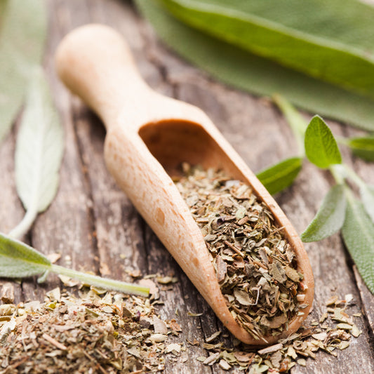 Dried sage leaves sit in a wooden scoop with loose sage scattered around it and fresh green sage leaves in the background.