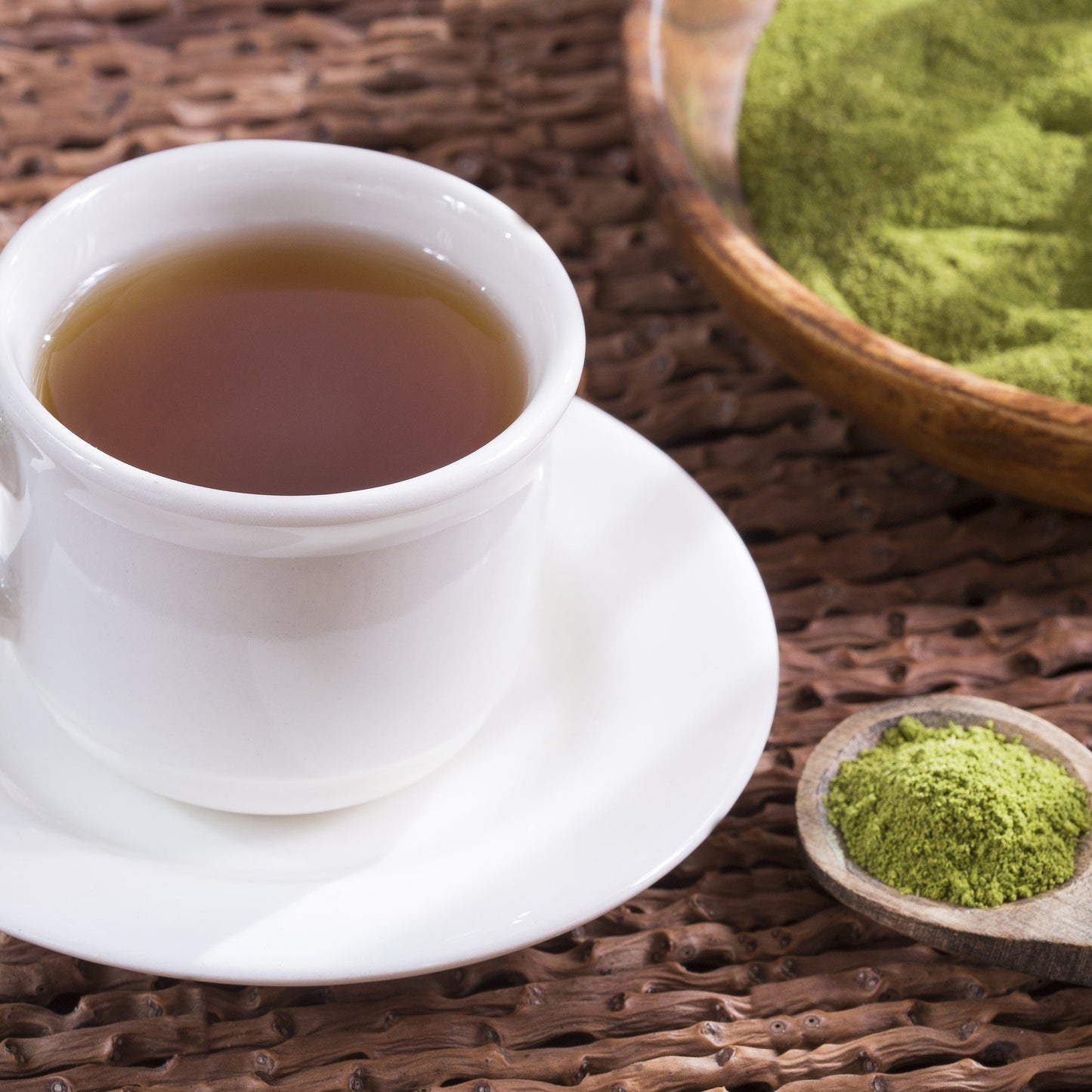 A white cup of brewed moringa drink sits on a saucer beside a bowl and spoon containing green moringa leaf powder on a rustic wooden surface.