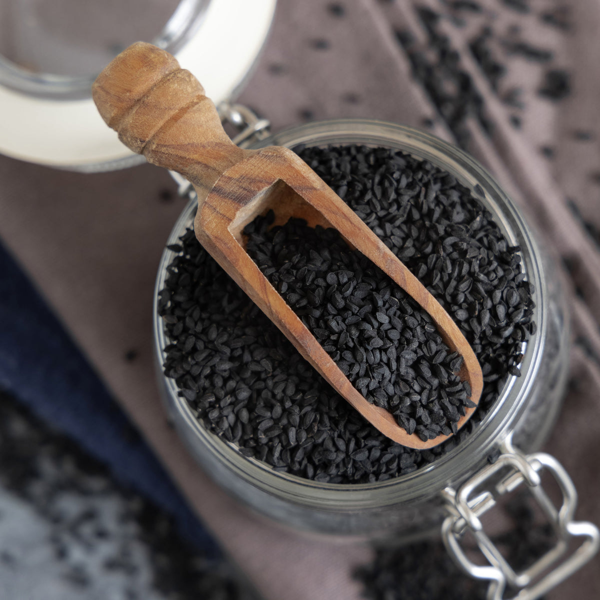 Organic nigella seeds fill a clear glass jar with a small wooden scoop resting across the top, showing the tiny black seeds and their naturally irregular texture.