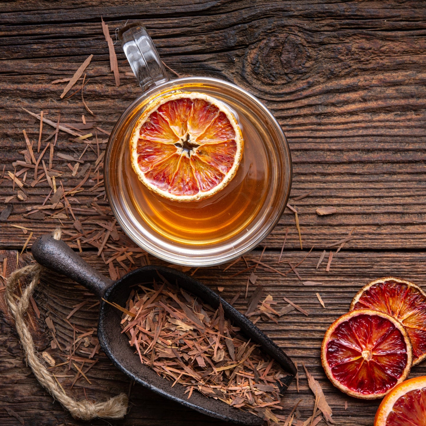A glass cup of brewed Pau D'Arco Bark Tea sits on a dark wooden surface with loose dried bark and dried citrus slices.
