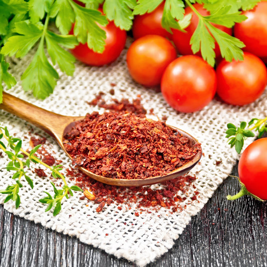 Dried tomato flakes are arranged on a rustic surface beside fresh red tomatoes and green herbs, showing the rich red colour and texture of the flakes.