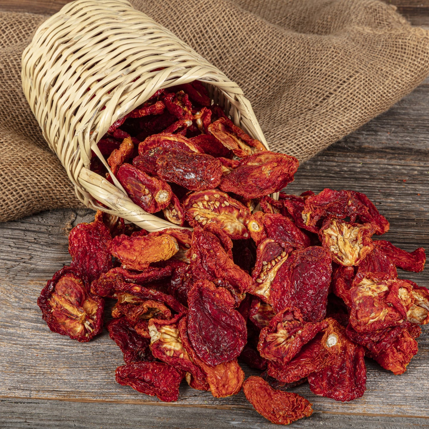 Dried red tomatoes spill from a small rustic bag onto a wooden surface, showing the deep red colour and irregular texture of the dried tomato pieces.