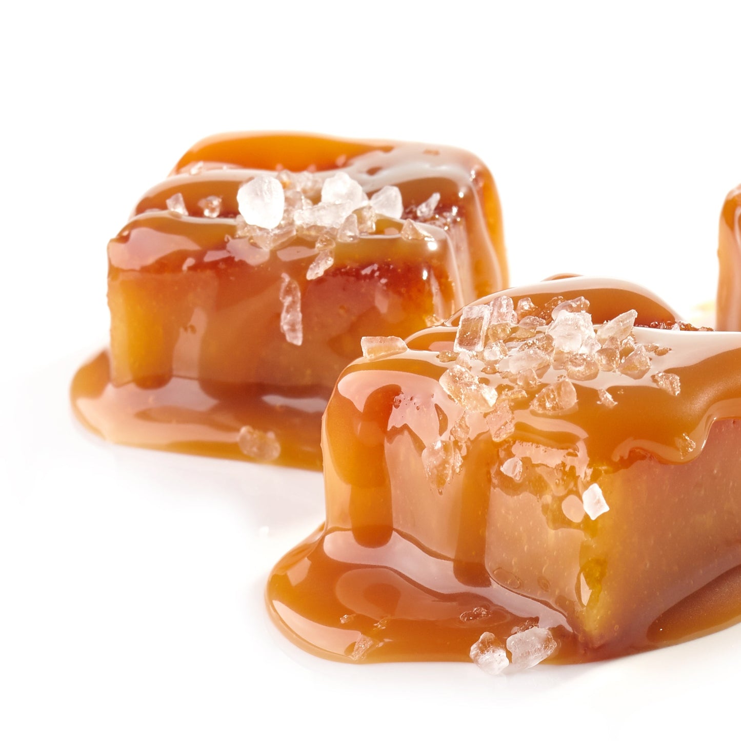 Pieces of golden salted caramel are stacked on a white surface with glossy caramel sauce and visible sea salt flakes.
