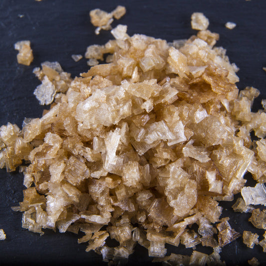A close-up pile of smoked sea salt flakes sits on a dark surface, showing the coarse, irregular texture and golden-brown colouring of the flakes.