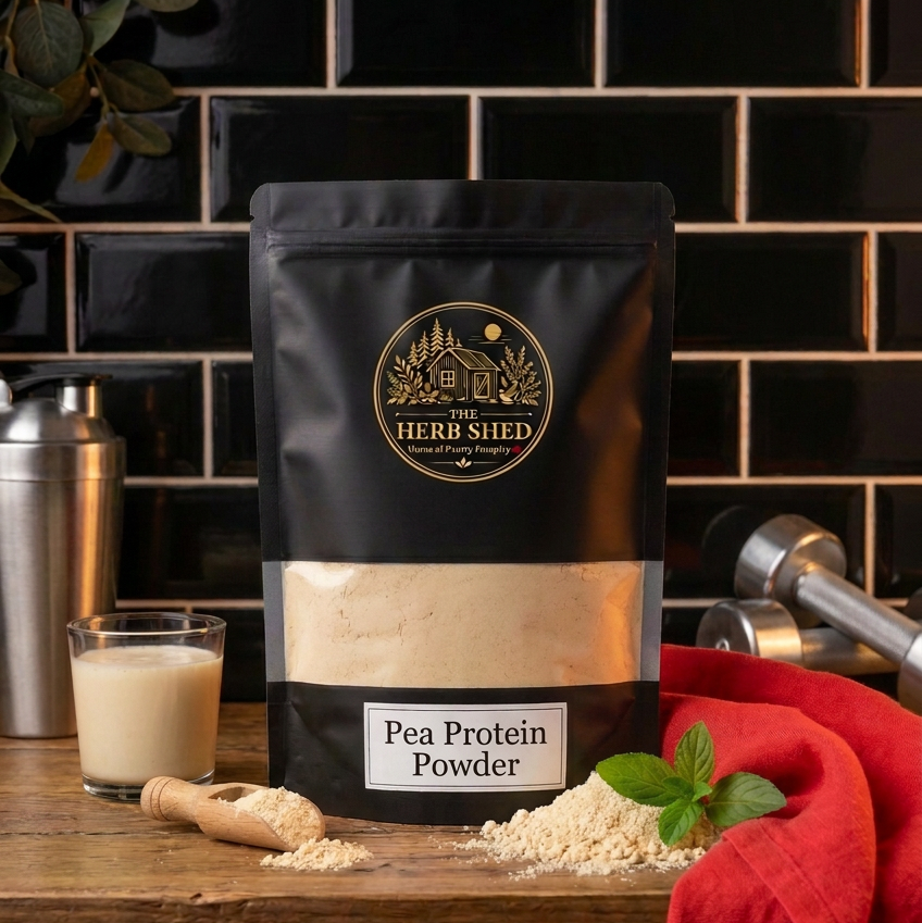A black stand-up pouch of The Herb Shed Pea Protein Powder in Wollongong, Australia, sits on a wooden surface with loose powder, a wooden scoop, shaker and glass, surrounded by fresh greenery and red fabric against a black tiled background.