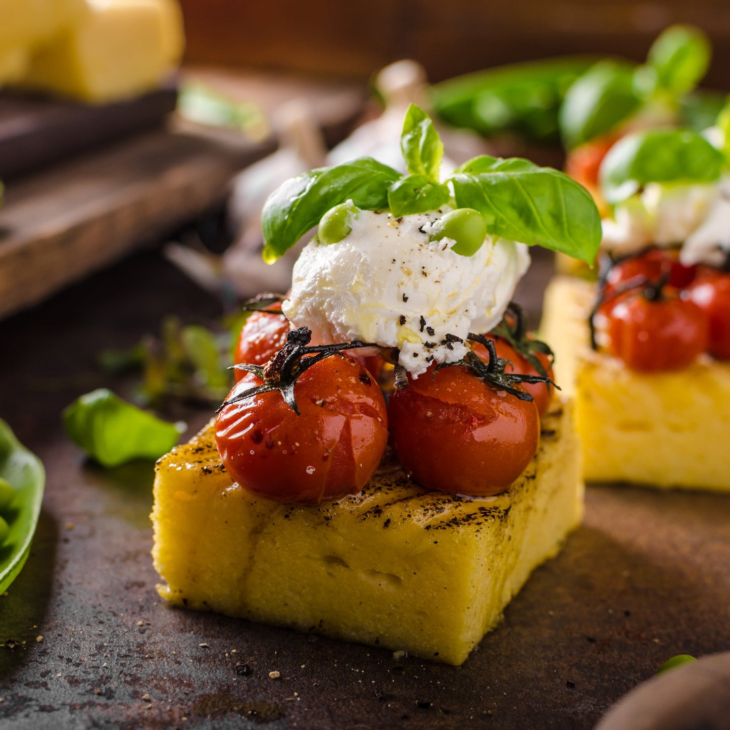 A square of cooked polenta is topped with roasted tomatoes, soft white cheese and fresh green herbs, showing a savoury serving idea for The Herb Shed Polenta Coarse.