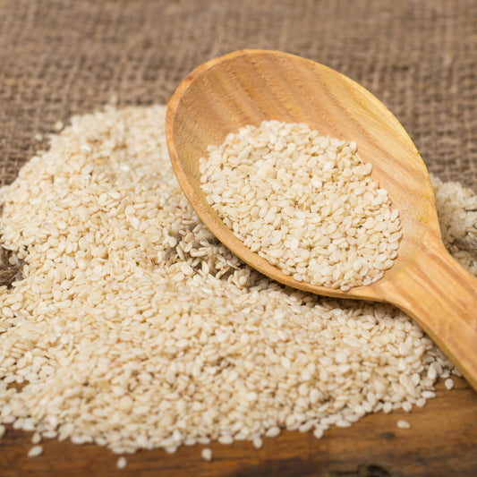 Roasted sesame seeds are scattered across a rustic surface with a wooden spoon resting among them, showing the small pale golden seeds and their toasted texture.
