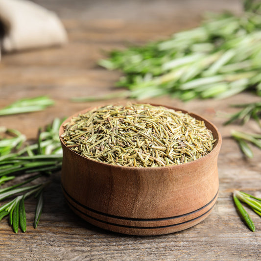 Dried Australian grown rosemary fills a small wooden bowl on a rustic timber surface, surrounded by fresh green rosemary sprigs and loose dried rosemary leaves.
