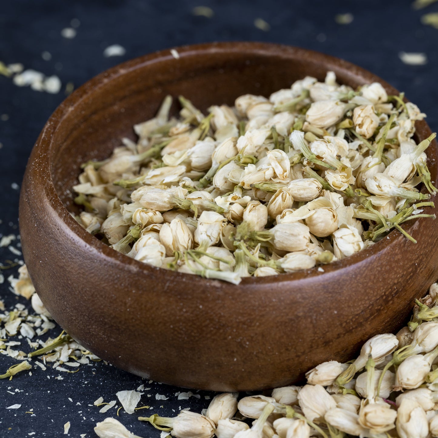 Jasmine flower buds in a brown wooden bowl on a dark blue table, with buds scattered and spilling out – delicate fragrant blossoms from The Herb Shed Wollongong