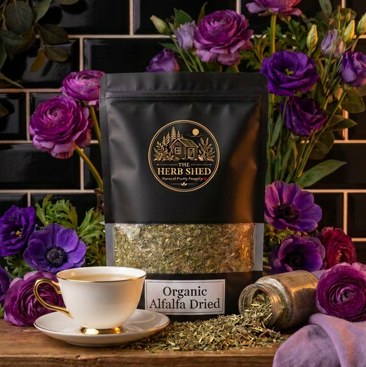 A black stand-up pouch of Herb Shied Organic Alfalfa Dried tea is displayed on a wooden surface with a white teacup, loose dried leaves, and a small jar, surrounded by purple flowers against a black tiled background.