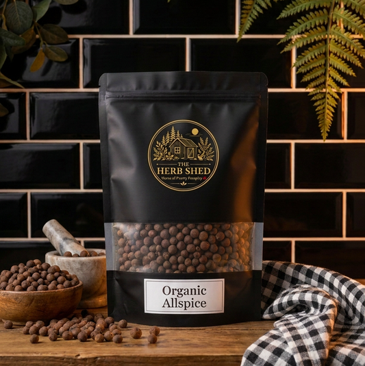 A stand-up pouch of Herb Shed Organic Allspice spice displayed on a rustic wooden counter against a black subway tile wall, with bowls of whole spices, a checkered cloth, and green foliage in the scene.