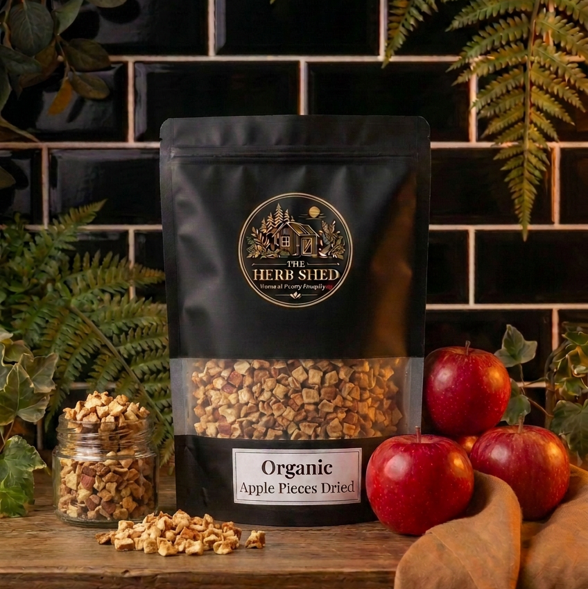 A black stand-up pouch of Herb Shed Organic Apple Pieces Dried, displayed on a rustic wooden surface with fresh red apples, a small glass jar of loose dried pieces, and green ferns against a black tiled background.