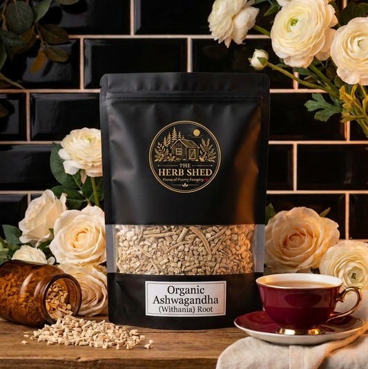 A black stand-up pouch of Heri Shed Organic Ashwagandha (Oshwagandha) Root sits on a wooden table, surrounded by white roses, a small jar of loose roots, and a red teacup on a saucer against a black tiled background.