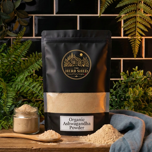 A black stand-up pouch of Herbi Shed Organic Ashwagandha Powder sits on a wooden surface with loose powder spilled around it, a small jar of powder, and a blue cloth. The background features a black tiled wall and green ferns.