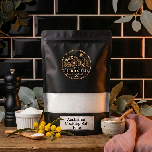 A black stand-up pouch of Heri Shied Australian Cooking Salt Salt, Fine, displayed on a rustic wooden table with a black tiled background, surrounded by kitchen props like a pepper mill, small bowls, and greenery.