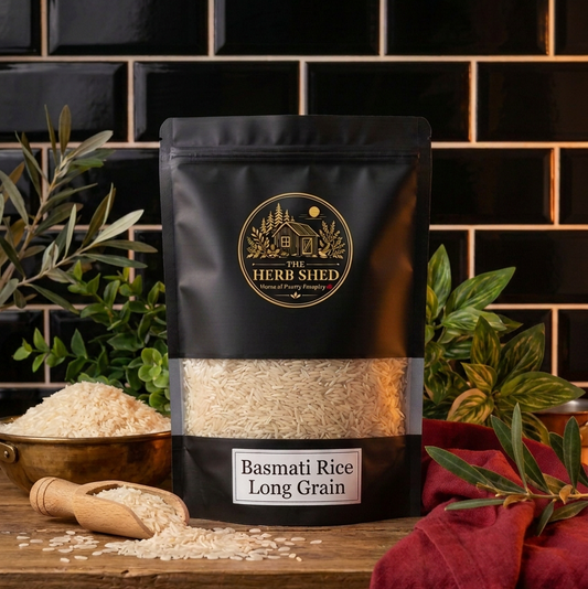 A black stand-up pouch of Herb Shed Basmati Rice Long Grain with a clear window showing the rice inside, styled on a wooden surface with a bowl of rice, a wooden scoop, and green plants against a black subway tile background.