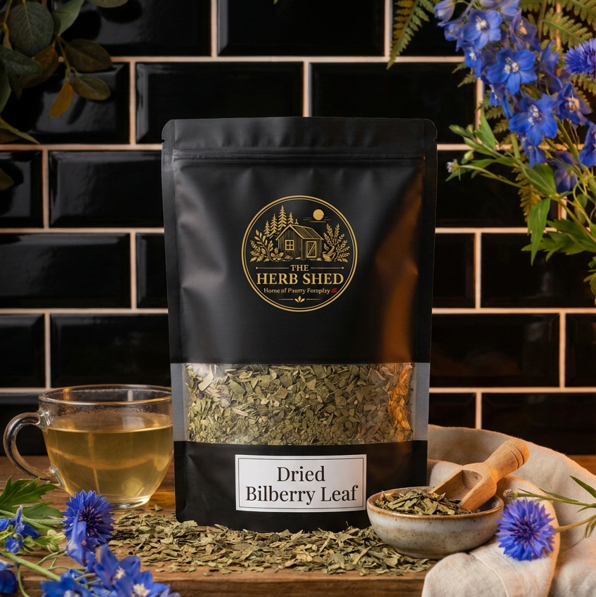 A stand-up pouch of Herb Shed Dried Bilberry Leaf tea placed on a wooden surface, surrounded by blue flowers, green leaves, a cup of tea, and a small bowl with a wooden scoop. The background is a black tiled wall.