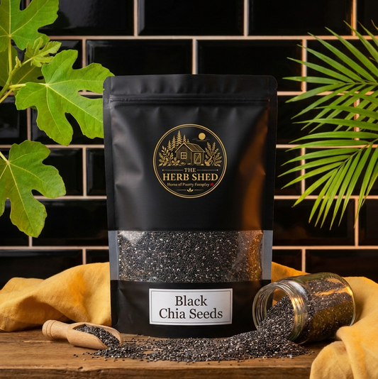 A black stand-up pouch of Heri Shed Black Chia Seeds with a clear window showing the seeds inside, styled on a wooden surface with a tipped-over jar, a wooden scoop, and yellow cloth against a black tiled background with green leaves.