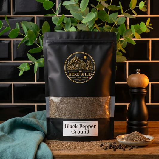 A black stand-up pouch of Heri Shed Ground Black Pepper sits on a wooden surface against a black tiled wall, styled with green eucalyptus leaves, a teal cloth, a pepper mill, and a small dish of loose pepper.