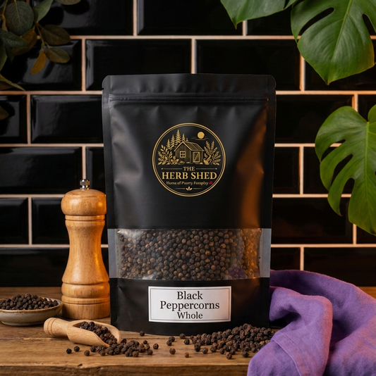 A black stand-up pouch of Herb Shed Black Peppercorns Whole, displayed on a rustic wooden table with scattered peppercorns, a wooden pepper mill, and a purple cloth, set against a black tiled wall with green leaves.