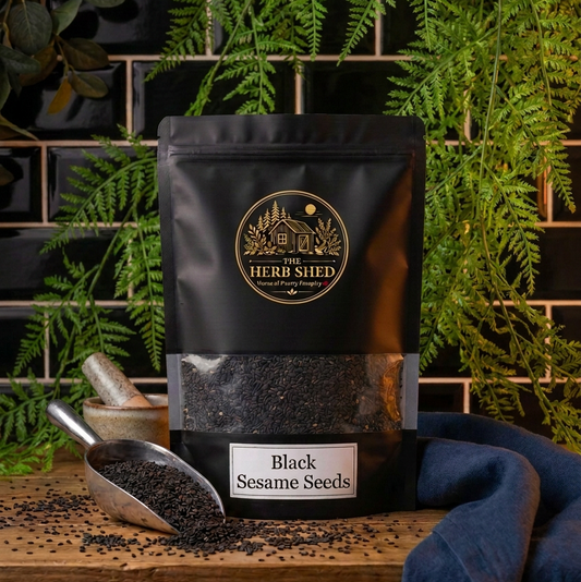 A black stand-up pouch of Herb Shiff Black Sesame Seeds sits on a wooden surface with scattered seeds, a metal scoop, and a blue cloth. Green ferns and dark tiled walls form the background.