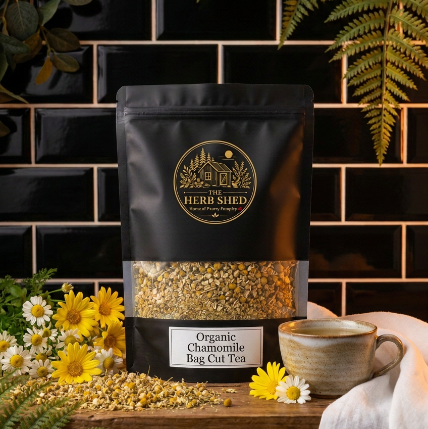 A stand-up pouch of Heri Shed Organic Chamomile Bag Cut Tea tea, displayed on a rustic wooden table with a ceramic mug, loose tea, and chamomile flowers against a black subway tile background.