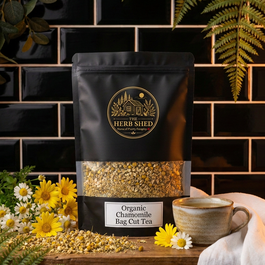 A stand-up pouch of Heri Shed Organic Chamomile Bag Cut Tea tea, displayed on a rustic wooden table with a ceramic mug, loose tea, and chamomile flowers against a black subway tile background.