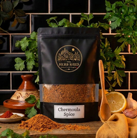 A black stand-up pouch of Herb Shed Chermoula Spice with a clear window showing the brown spice inside, styled on a wooden surface with parsley, a ceramic pot, a wooden spoon, garlic, and a lemon slice against a black tiled background.