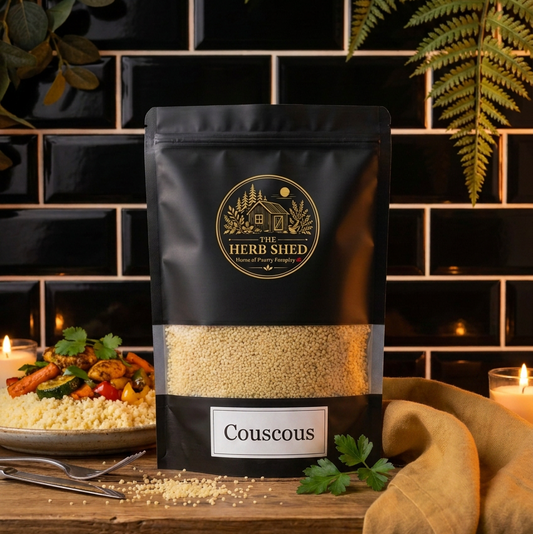 A stand-up pouch of Herbs Shed Couscous placed on a wooden table in a styled kitchen-like setting with black subway tiles, greenery, a plate of prepared couscous, and a candle.