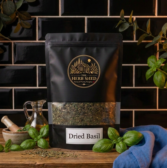 A black stand-up pouch of Herbs Shed Dried Basil sits on a wooden shelf against a black tiled wall, surrounded by fresh green basil leaves, a small glass bottle, and a blue cloth.