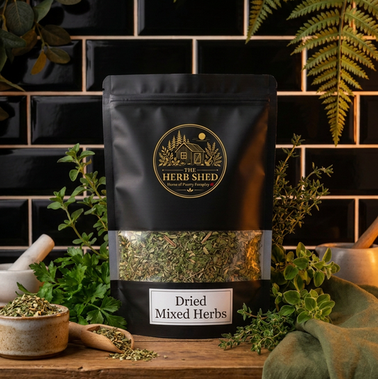 A black stand-up pouch of Herb Shed Dried Mixed Herbs sits on a wooden surface against a black tiled wall, surrounded by fresh green herbs, a mortar and pestle, and a green cloth.