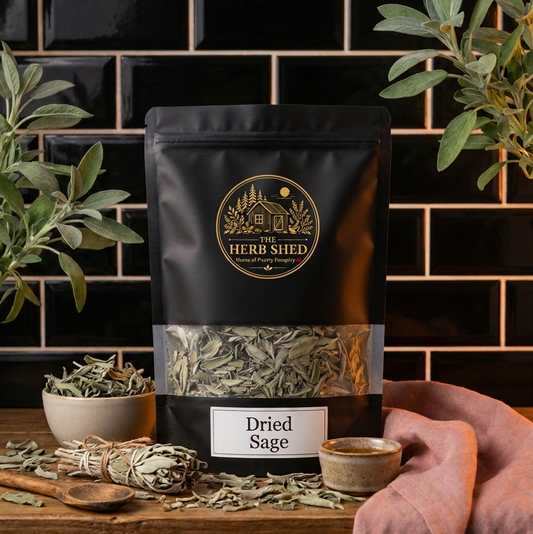 A black stand-up pouch of Herb Shed Dried Sage stands on a wooden surface against a glossy black subway tile wall, surrounded by olive branches, a small bowl of loose herbs, a sage bundle, and a pink cloth.