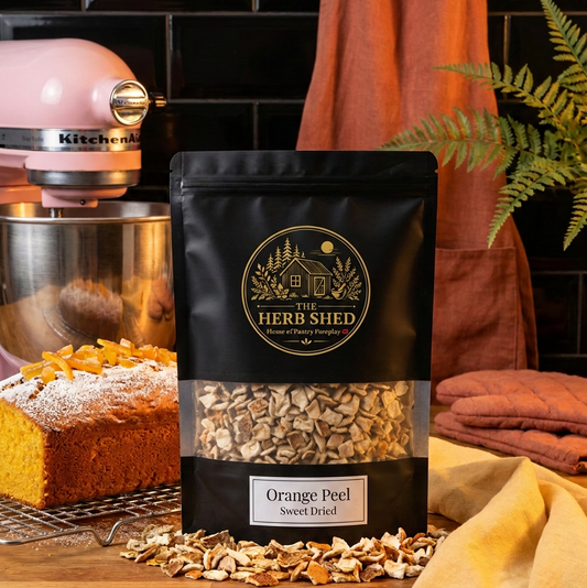 A black stand-up pouch of Herb Shed Orange Peel Sweet Dried Orange Peels stands on a wooden countertop, surrounded by scattered orange peel pieces. The scene includes a pink KitchenAid mixer, a loaf of bread, a terracotta apron, and a yellow cloth against a black tiled background.