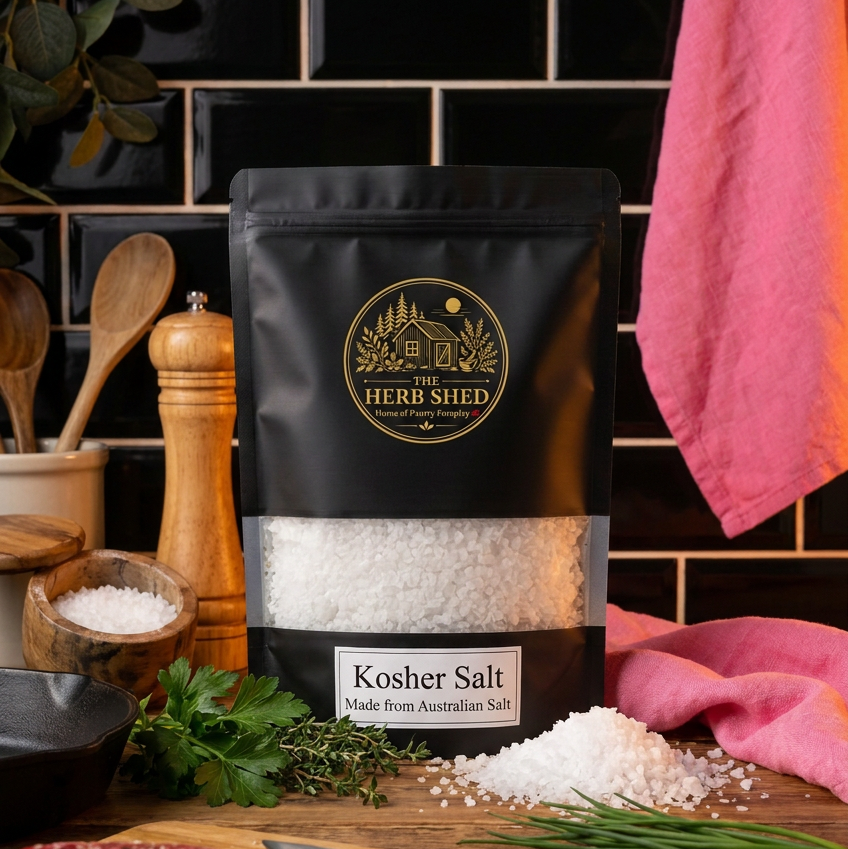 A black stand-up pouch of Herb Shed Kosher Salt stands on a wooden cutting board with a raw steak, knife, chives, parsley, and a pepper mill. The background features a black subway tile wall and a pink towel.