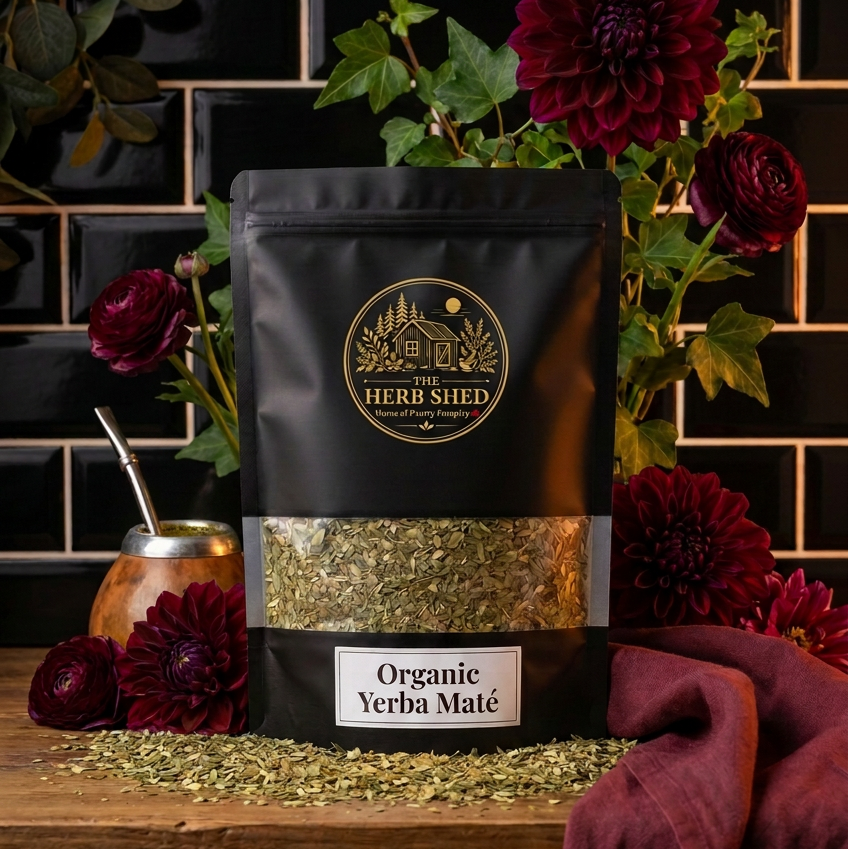 A black stand-up pouch of Herb Shed Organic Yerba Maté sits on a wooden surface with loose green tea scattered in front. 