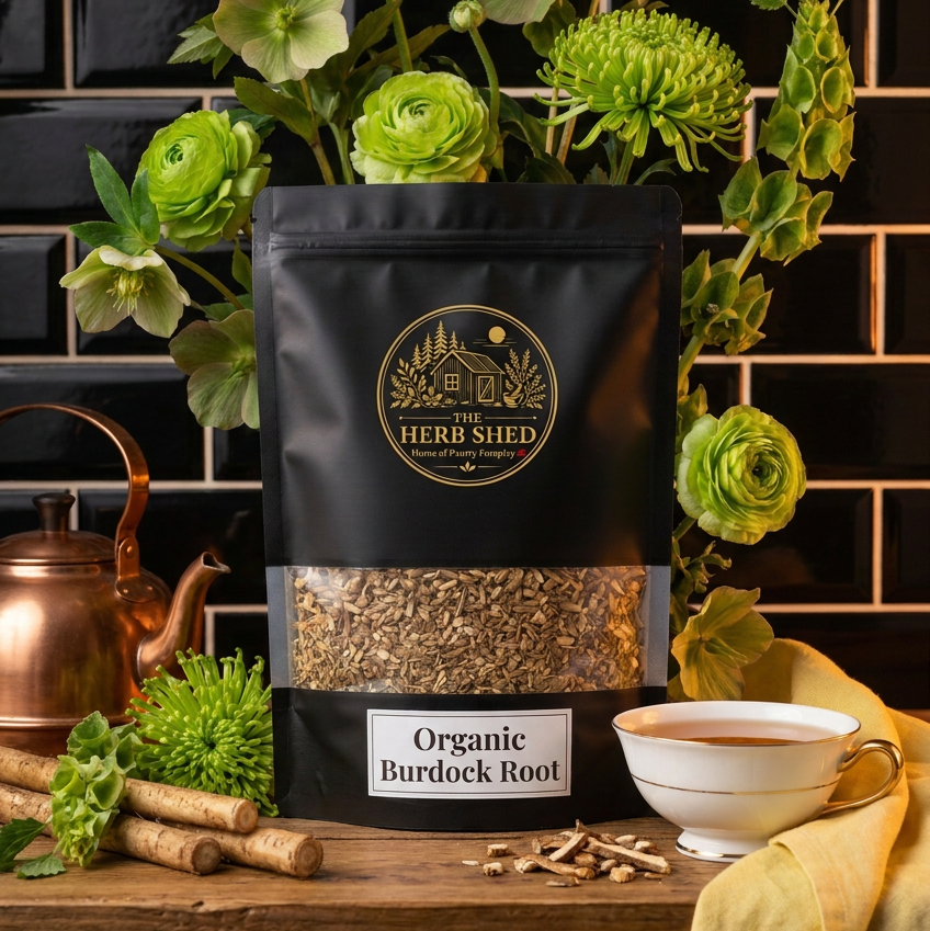 A black stand-up pouch of Herb Shed Organic Burdock Root tea sits on a wooden table surrounded by green flowers, cumin, a copper teapot, and a white teacup against a black tiled wall.