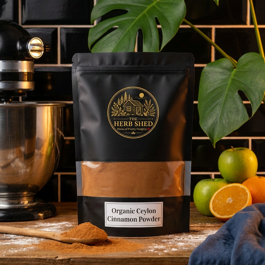 A black stand-up pouch of Herb Shed Organic Ceylon Cinnamon Powder on a rustic wooden counter, styled with a metal stand mixer, a wooden spoon of cinnamon powder, green and orange fruits, and a blue cloth against a black tiled background.