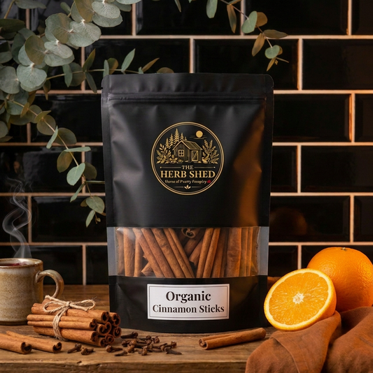 A black stand-up pouch of Herb Shed Organic Cinnamon Sticks stands on a wooden surface with cinnamon sticks, cloves, a mug with steam, eucalyptus leaves, and orange slices around it against a black tiled wall.